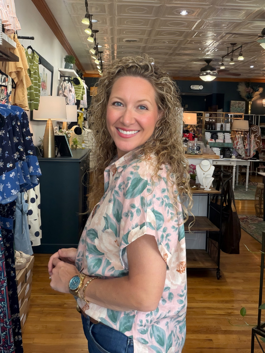 Woman standing in a clothing store wearing a floral shirt, smiling at the camera.