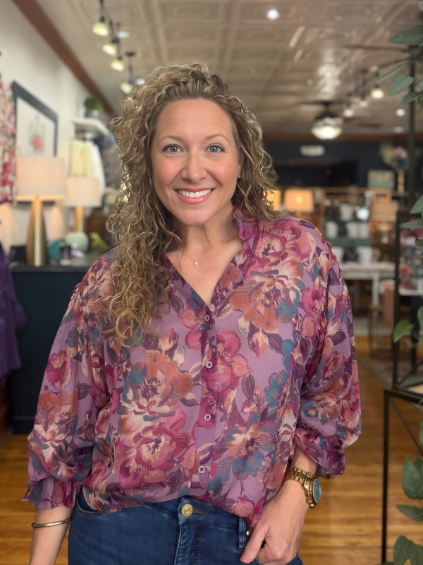 A smiling woman with curly blonde hair wearing a purple and pink floral button-up blouse with blue jeans, standing inside a retail store.