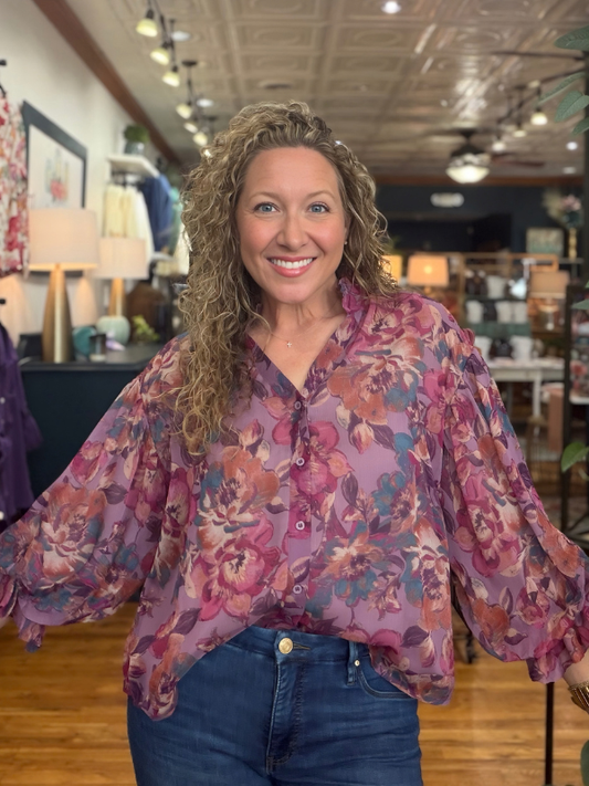 A smiling woman with curly blonde hair wearing a sheer purple floral blouse with blue jeans, standing inside a retail store.