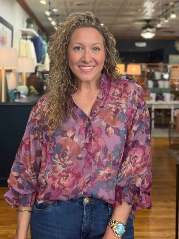 A smiling woman with curly blonde hair wearing a purple floral button-down blouse and blue jeans, standing in a retail store.