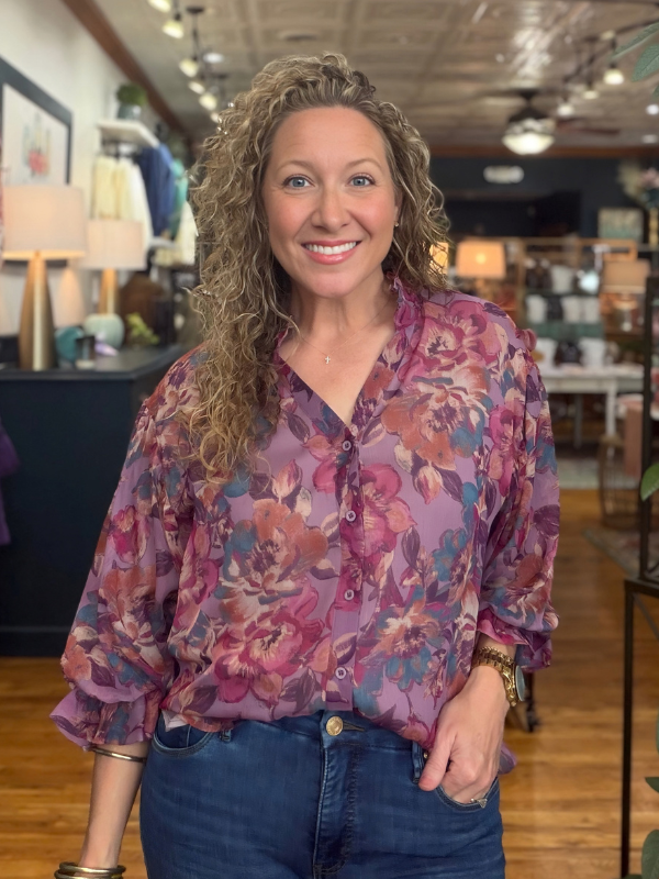 A smiling woman with curly blonde hair wearing a purple floral button-down blouse and blue jeans, standing inside a retail store.