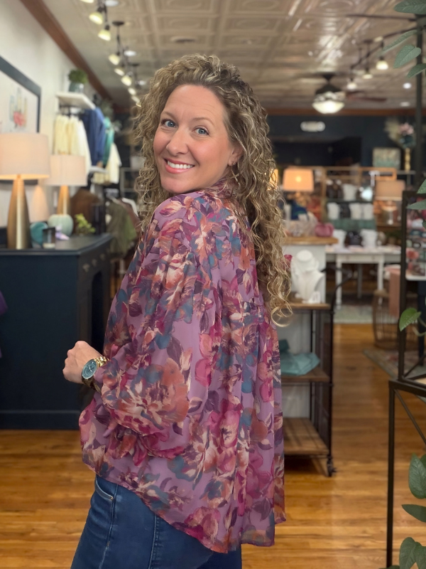 A woman with curly blonde hair wearing a purple and pink floral long-sleeve blouse and blue jeans, posing inside a boutique store.