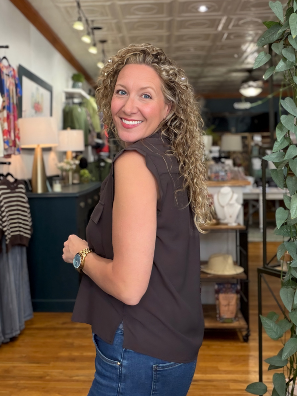 A woman with curly blonde hair wearing a sleeveless dark brown top and blue jeans, smiling and looking back in a retail store setting.
