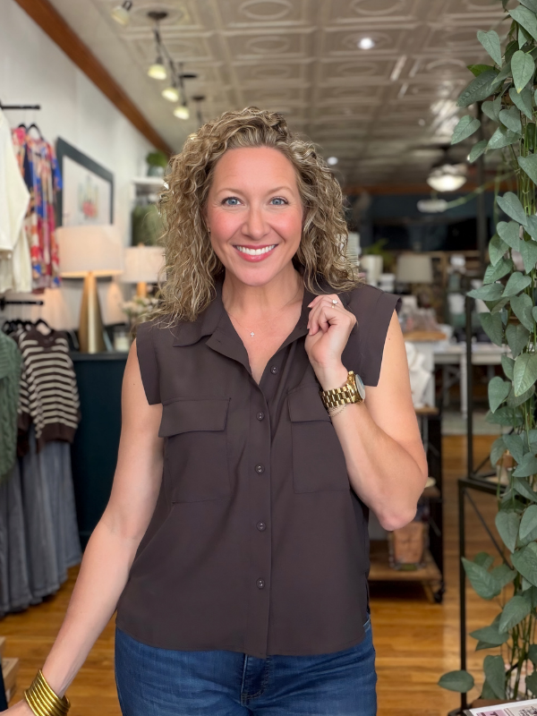 A smiling woman with curly blonde hair wearing a sleeveless dark brown button-down shirt and blue jeans, standing inside a boutique store.