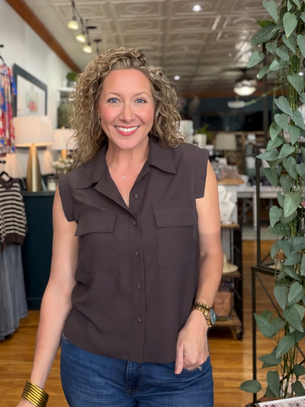A smiling woman with curly blonde hair wearing a sleeveless dark brown button-up shirt and blue jeans, standing inside a retail store with shelves, lamps, and a plant visible in the background.