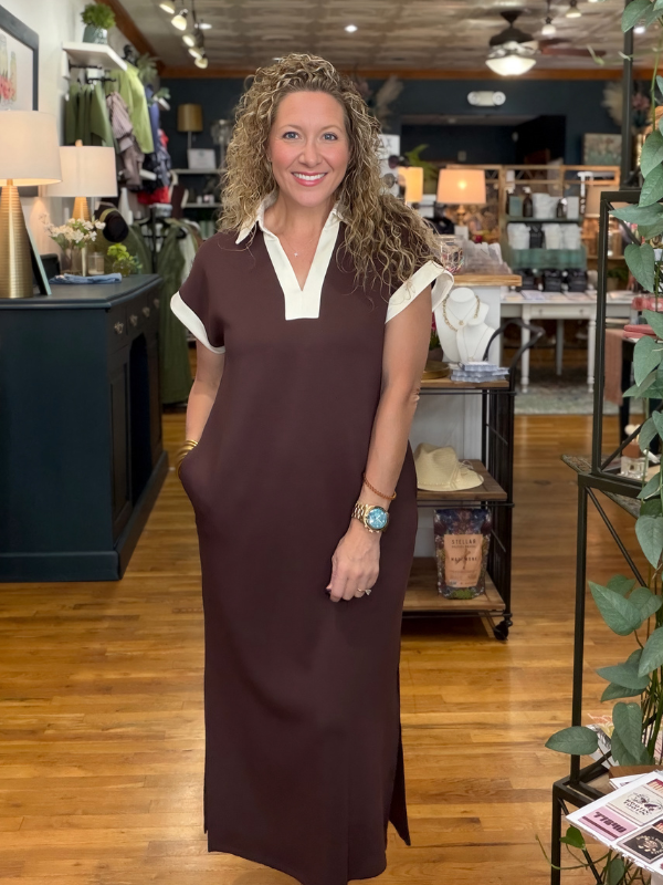 A woman with curly hair stands in a retail store wearing a dark brown maxi dress with a white collar and white sleeve cuffs. The background shows a busy boutique interior with wooden floors, shelves, and other merchandise.