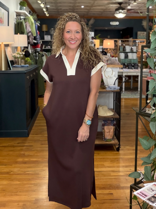 A woman with curly hair stands in a retail store wearing a dark brown maxi dress with a white collar and white sleeve cuffs. The background shows a busy boutique interior with wooden floors, shelves, and other merchandise.
