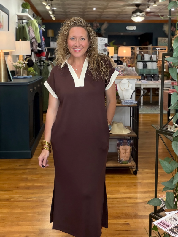 A woman with curly blonde hair wearing a brown maxi dress with cream-colored collar and sleeve trim, standing inside a retail store with wooden floors and home decor items in the background.