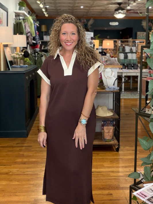 A woman with curly brown hair stands in a boutique wearing a dark brown short-sleeve maxi dress with a contrasting white collar and sleeve trim.