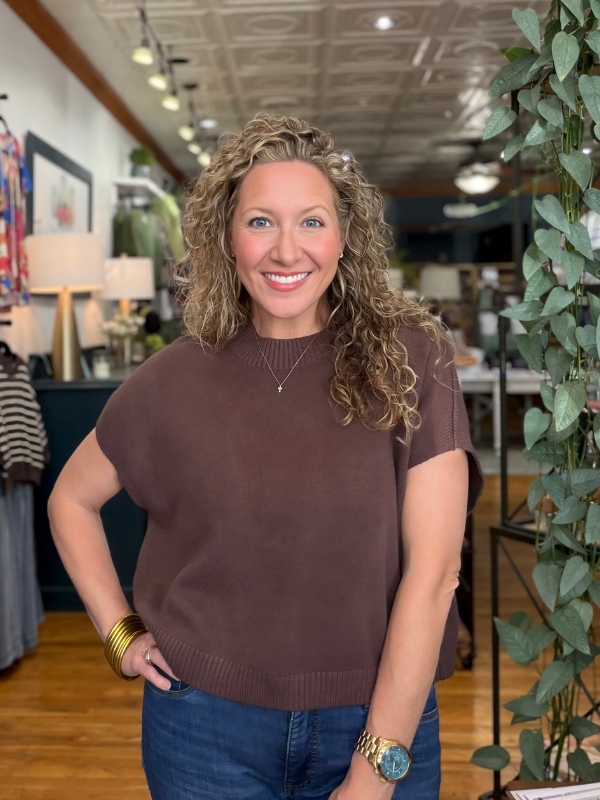 A smiling woman with curly blonde hair wearing a brown short-sleeve knit top and blue jeans, standing in a boutique interior with plants and store fixtures visible in the background.
