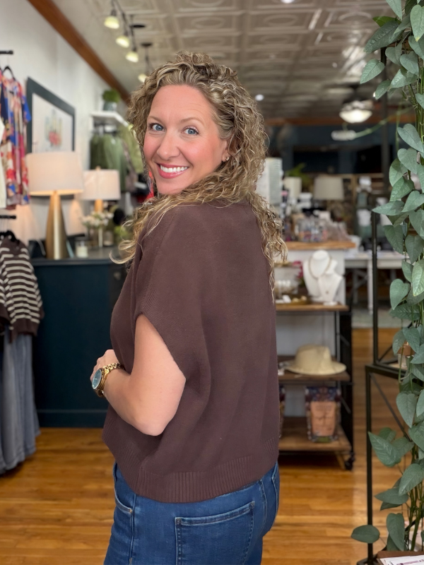 A woman with curly blonde hair is shown from the back in a boutique, wearing a brown short-sleeve knit top and blue jeans.