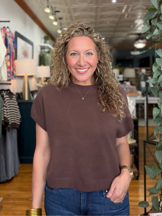 A smiling woman with curly blonde hair wearing a short-sleeve brown knit sweater and blue jeans, standing in a retail store interior.