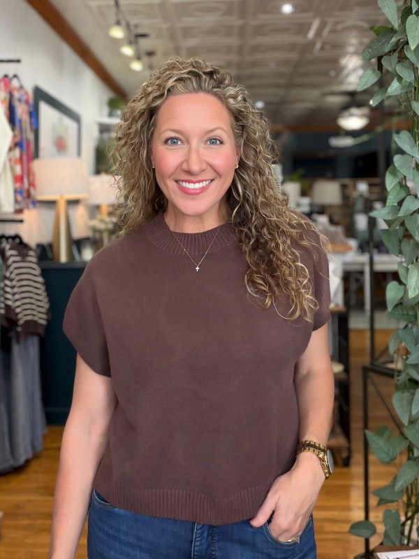 A smiling woman with curly blonde hair wearing a short-sleeve brown knit sweater and blue jeans, standing in a retail store interior with clothing racks, lamps, and a plant visible in the background.