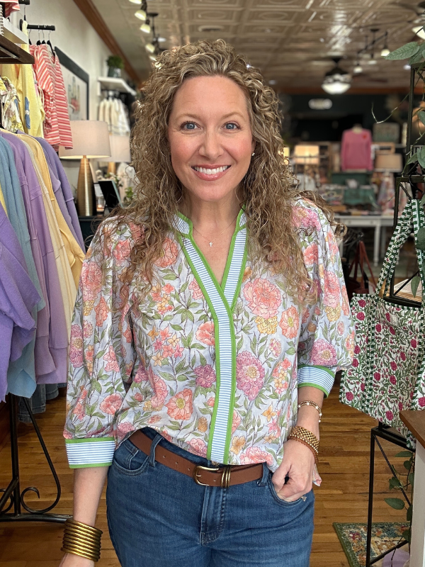 Woman in a floral blouse and jeans standing in a store with clothing racks in the background.