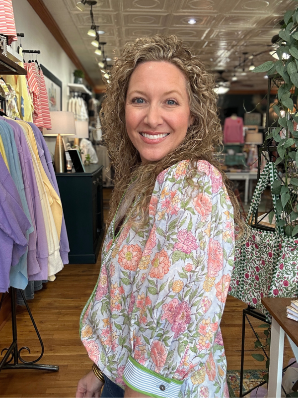 Woman in a floral blouse standing in a store with clothing and decor around her.