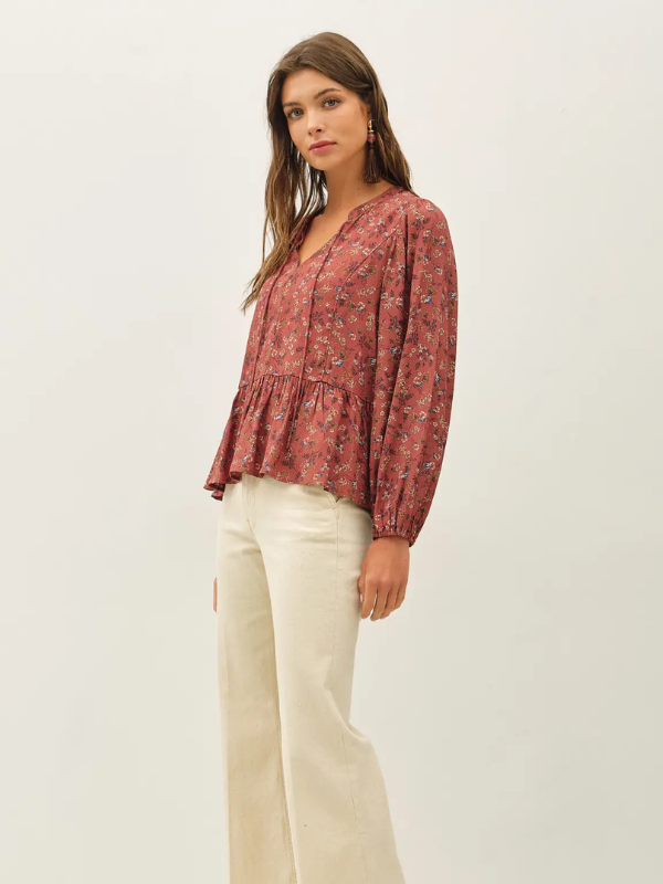 A woman modeling a long-sleeve, tiered peplum blouse with a rust-red and blue floral pattern, paired with cream-colored wide-leg trousers, against a plain off-white studio background.