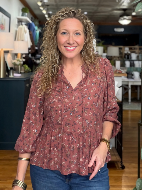 A smiling woman with curly blonde hair wearing a dusty rose long-sleeve blouse with a small floral print and blue jeans, standing inside a boutique.