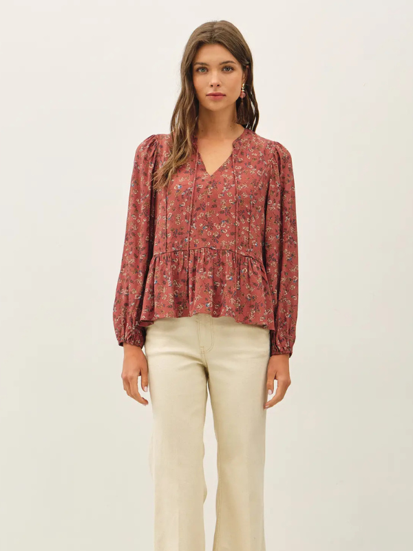 A woman modeling a long-sleeve, V-neck blouse with a dusty rose and red floral pattern, paired with cream-colored pants, against a plain off-white background.