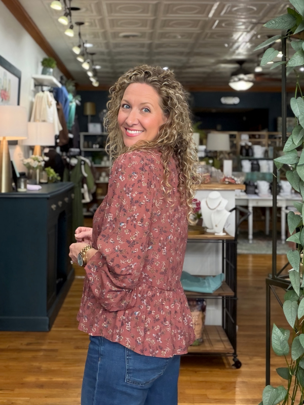A woman with curly blonde hair wearing a dusty rose floral long-sleeve blouse and blue jeans, posing inside a boutique store.