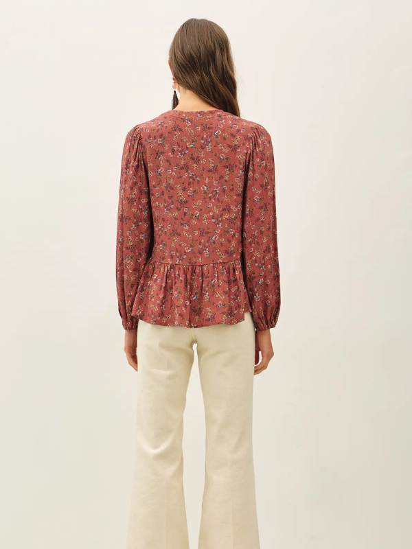 A woman viewed from the back wearing a long-sleeve floral blouse in a muted terracotta red with small blue and green floral details, paired with cream-colored pants, against a plain off-white background.