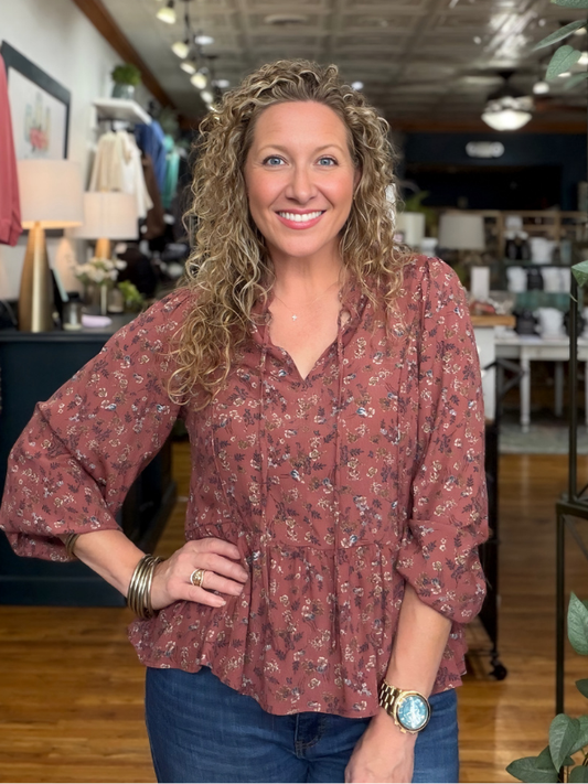 A smiling woman with curly blonde hair wearing a dusty rose long-sleeve button-up blouse with a small floral print and blue jeans, standing inside a boutique.