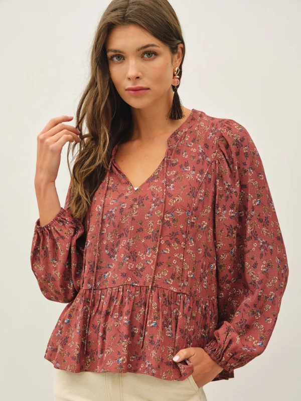 A woman wearing a dusty rose long-sleeve blouse with a dense small floral print, featuring a split V-neckline and a gathered peplum hem, paired with cream pants against a light gray studio background.