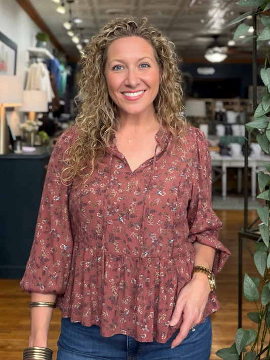 A smiling woman with curly blonde hair wearing a rust-colored, long-sleeve button-up blouse with a small floral print and blue jeans, standing in a retail store.