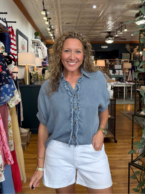 Woman standing in a store wearing a blue shirt and white shorts, surrounded by clothing and decor.
