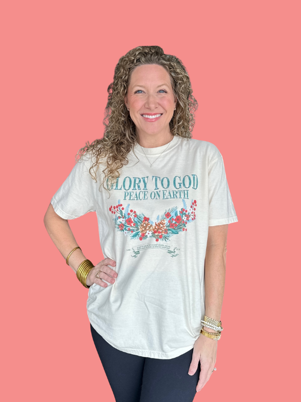 Woman wearing a white t-shirt with 'Glory to God Peace on Earth' text and floral design on a pink background