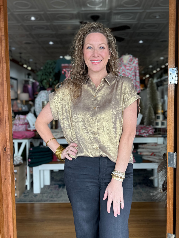 Woman in a gold top and dark pants standing in a store with various items in the background.