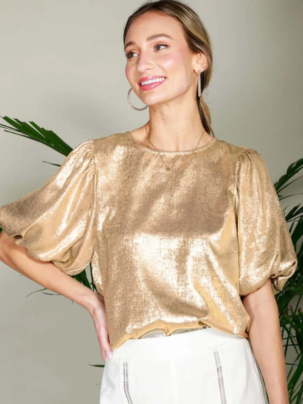Woman wearing a gold metallic top with white pants against a neutral background