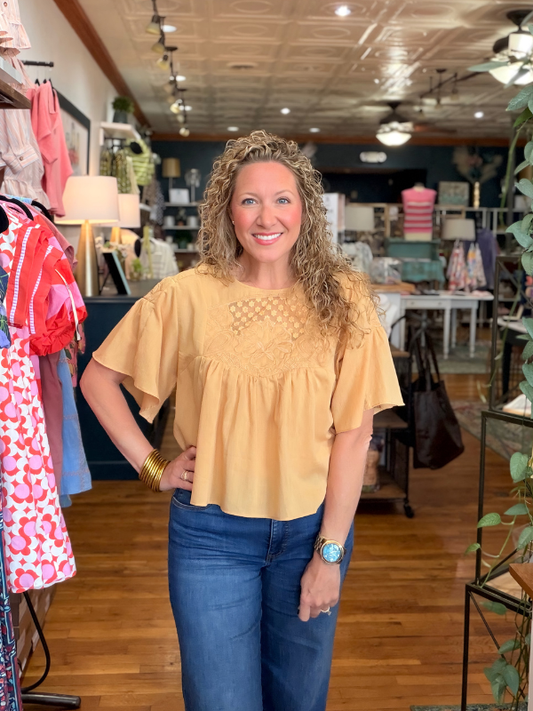 Woman in a yellow top and blue jeans standing in a store with clothing racks and products in the background.