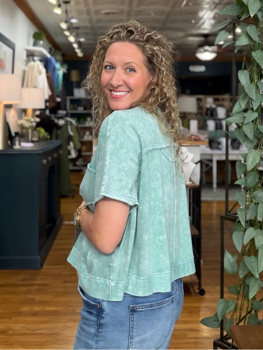 A woman with curly blonde hair wearing a light mint green washed short-sleeve crop top and blue jeans, standing in a retail store.