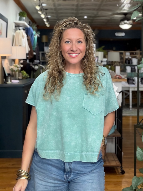 A smiling woman with curly blonde hair wearing a light mint green acid-wash short-sleeve t-shirt with a chest pocket, paired with blue jeans, standing inside a boutique store.