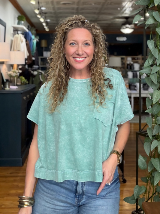 A smiling woman with curly blonde hair wearing a light mint green washed short-sleeve t-shirt and blue jeans, standing inside a retail store.
