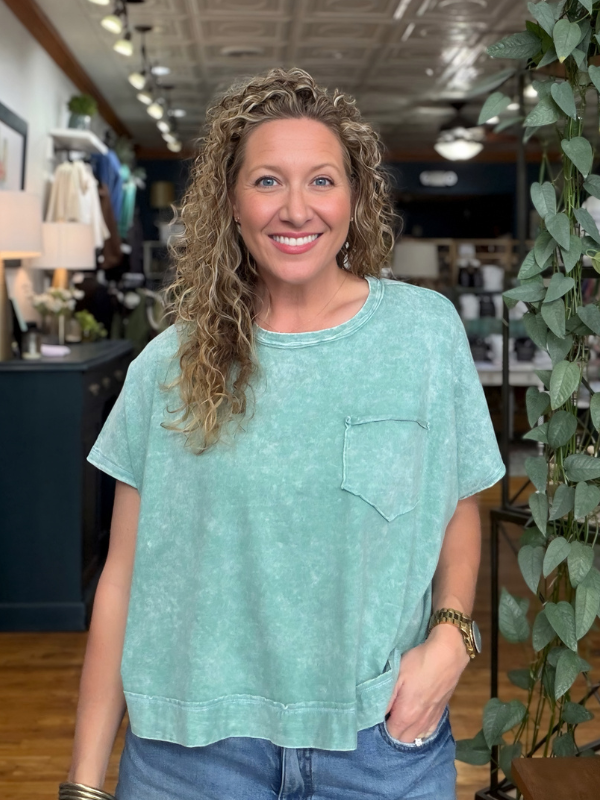 A smiling woman with curly blonde hair wearing a light mint green acid-wash short-sleeve t-shirt with a chest pocket, paired with blue jeans, standing inside a boutique store.
