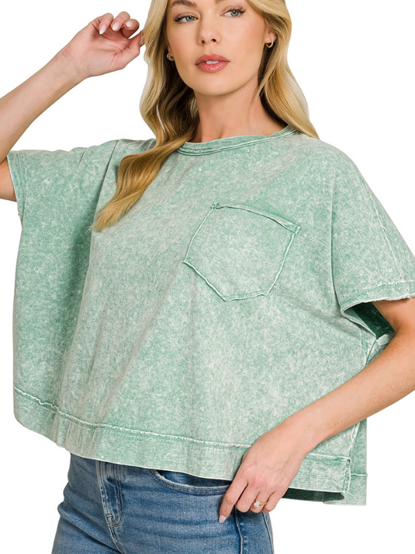 A woman wearing a light green acid-wash short-sleeve crop top with a chest pocket, paired with blue jeans, against a plain white background.