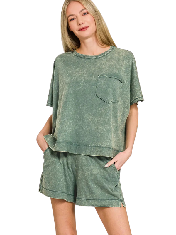 A woman modeling a matching two-piece lounge set in a washed sage green color, featuring a short-sleeve cropped top with a chest pocket and matching shorts, set against a plain white background.
