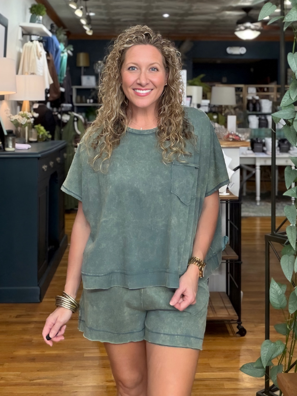 A smiling woman with curly blonde hair stands in a boutique wearing a matching sage green short-sleeve top and shorts set. The photo is taken in a retail store with wood floors, shelves, and plants in the background.