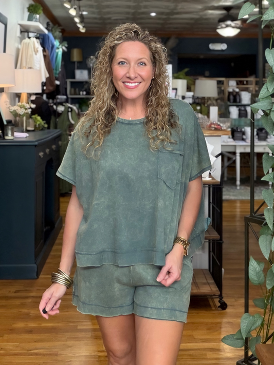 A smiling woman with curly blonde hair stands in a boutique wearing a matching sage green short-sleeve top and shorts set. The photo is taken in a retail store with wood floors, shelves, and plants in the background.