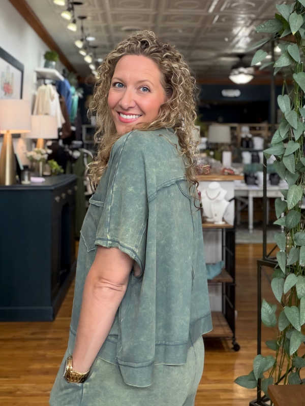 A smiling woman with curly blonde hair stands in a boutique wearing a matching sage green short-sleeve top and shorts set. The set has a washed, textured finish and includes a chest pocket on the top.