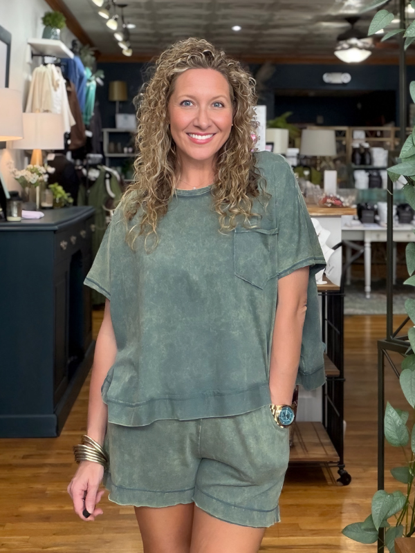 A smiling woman with curly blonde hair stands in a boutique wearing a matching sage green short-sleeve top and shorts set. The set has a washed, textured finish and includes a chest pocket on the top.