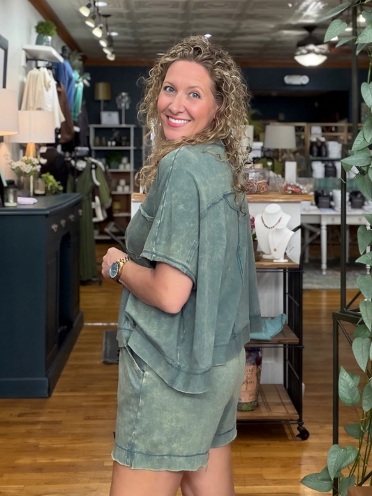 A woman with curly blonde hair stands in a boutique shop, wearing a matching sage green short-sleeve top and shorts set. She is looking back over her shoulder in a store with wooden floors, clothing racks, and shelves in the background.