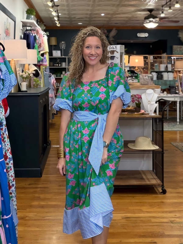 Woman in a floral dress standing in a store with various items on shelves and tables.