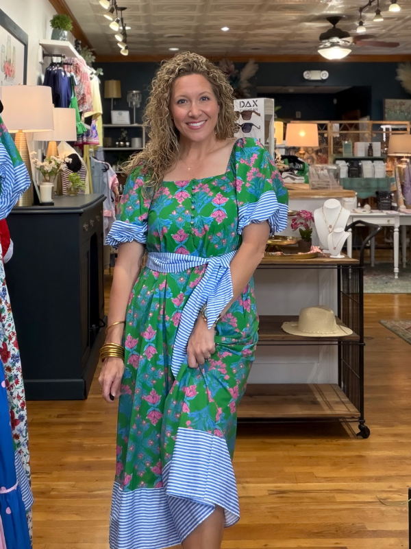 Woman in a floral dress standing in a store with various items on shelves and tables.