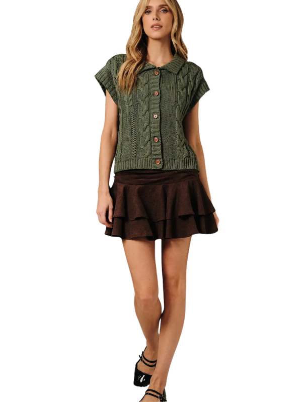 A woman modeling a short-sleeve olive green cable-knit button-front top paired with a dark brown ruffled mini skirt and black strappy heels, set against a plain white background.