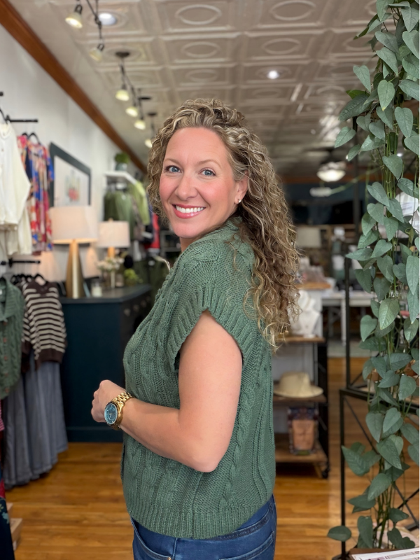 A smiling woman with curly blonde hair wearing a green cable-knit sleeveless sweater vest and blue jeans, standing inside a boutique store.