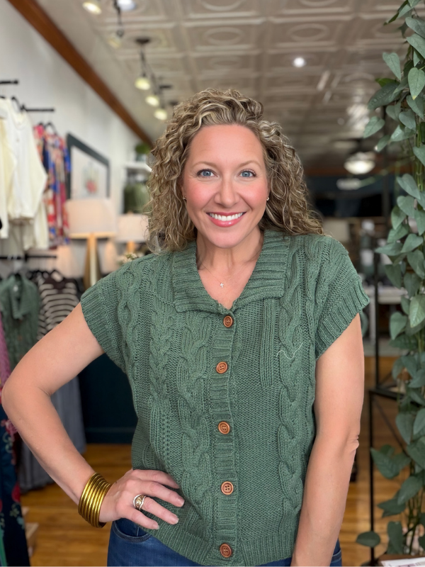 A smiling woman with curly blonde hair wearing a sage green cable-knit short-sleeve button-up sweater, standing inside a clothing boutique.