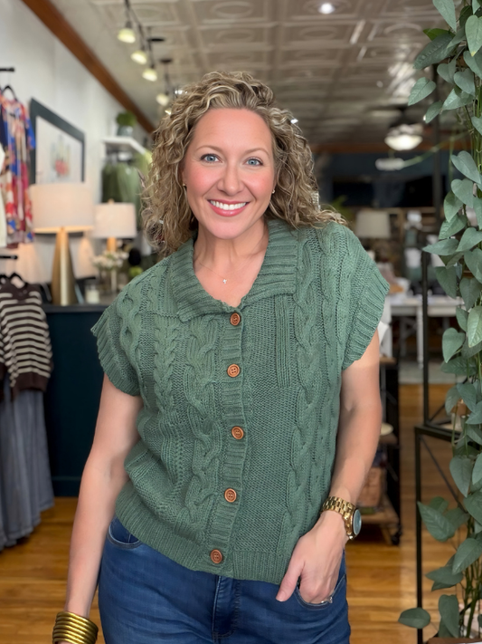 A smiling woman with curly blonde hair wearing a sage green cable-knit short-sleeve button-up sweater and blue jeans, standing inside a retail store.