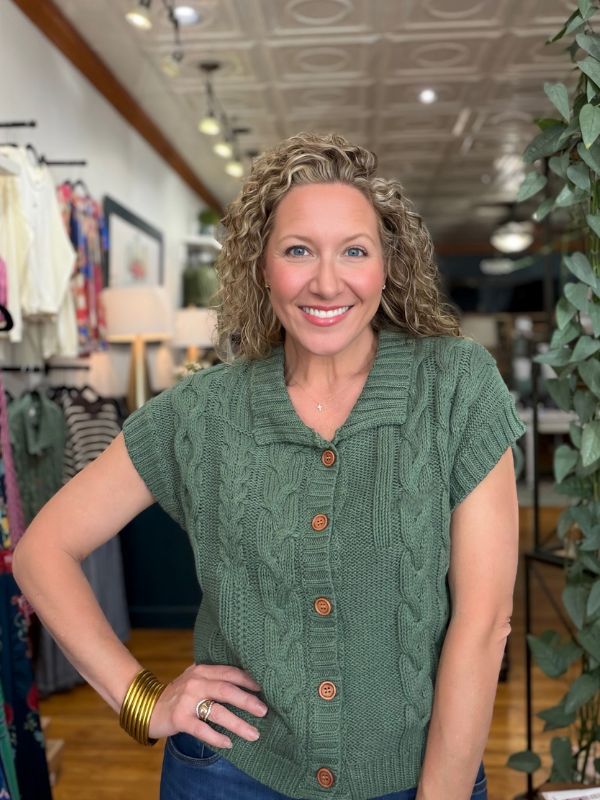 A smiling woman with curly blonde hair wearing a green cable-knit short-sleeve button-up sweater, standing in a boutique store.
